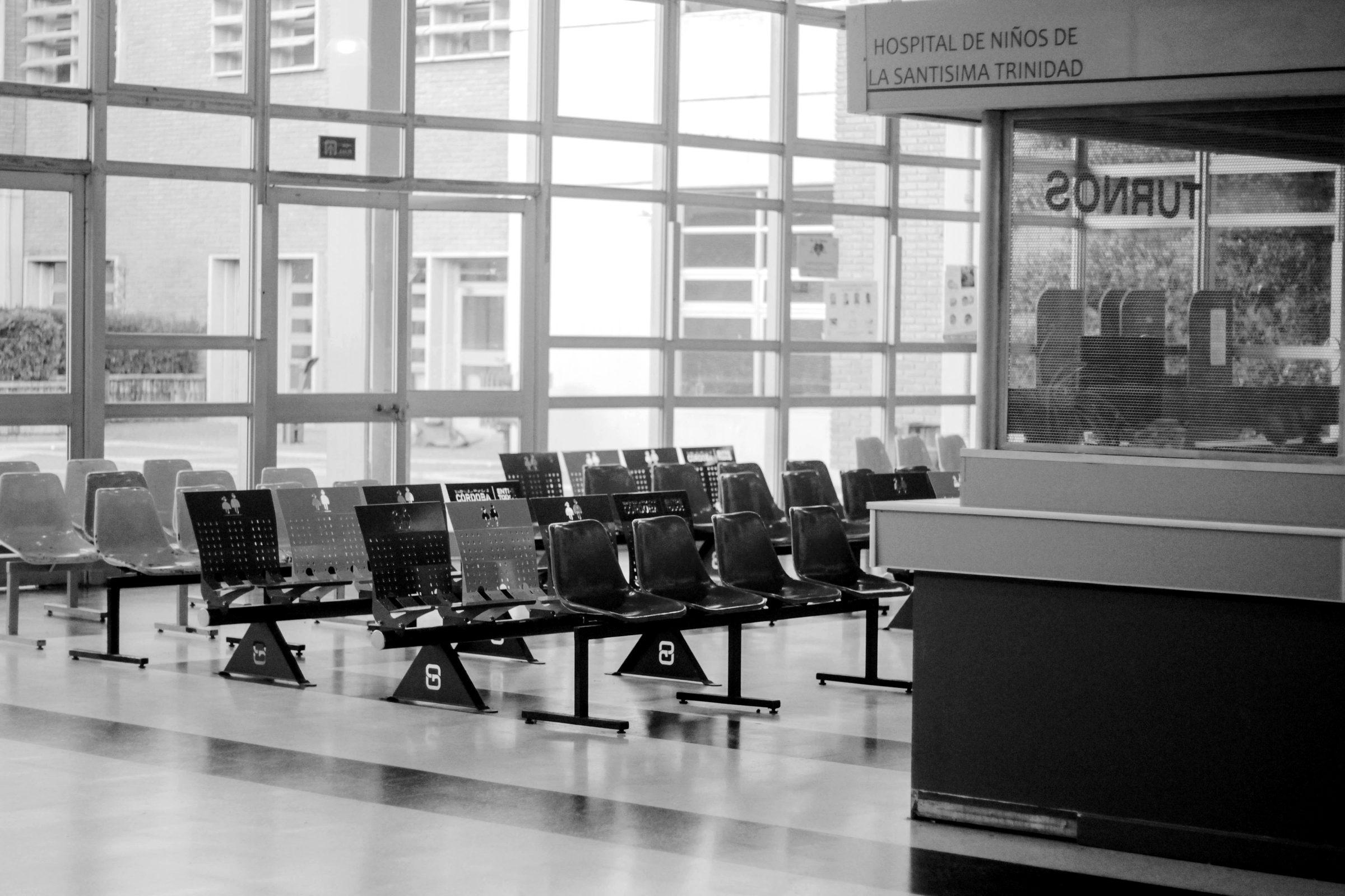 A hospital waiting room with rows of chairs and a numbered queue window